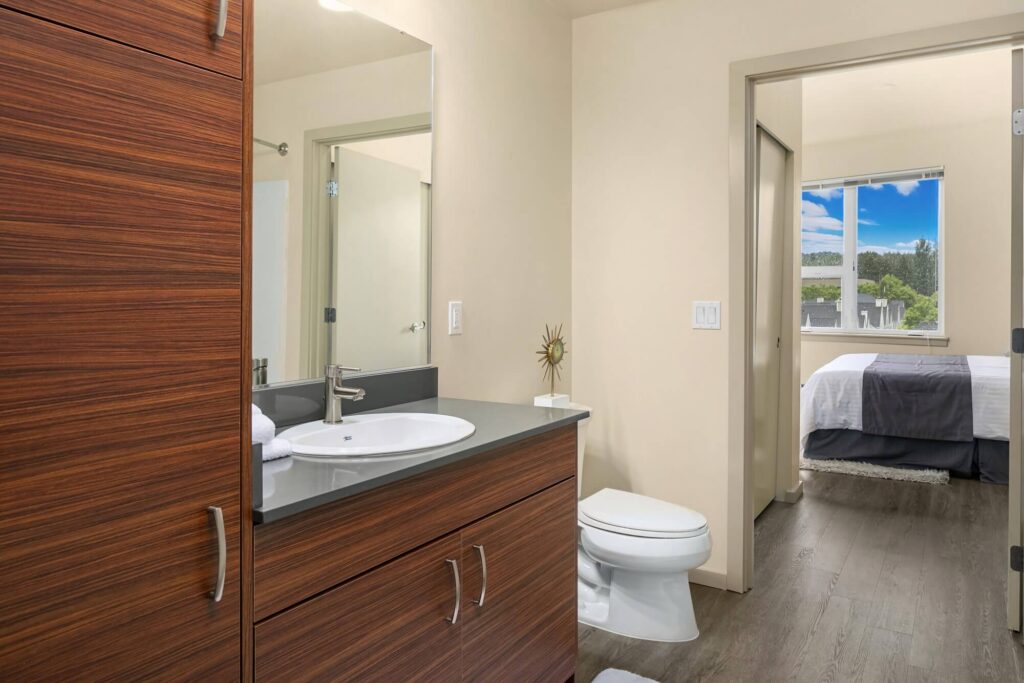 Modern bathroom with wood cabinets, white sink, and toilet; view extends into a bedroom with a large window showing a blue sky and greenery outside. The space features neutral colors and clean lines.