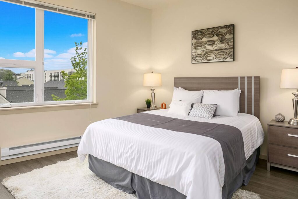 A modern bedroom with a large bed, white linens, gray throw, two nightstands with lamps, abstract wall art, a window with a view of buildings and trees, and light-colored walls and flooring.