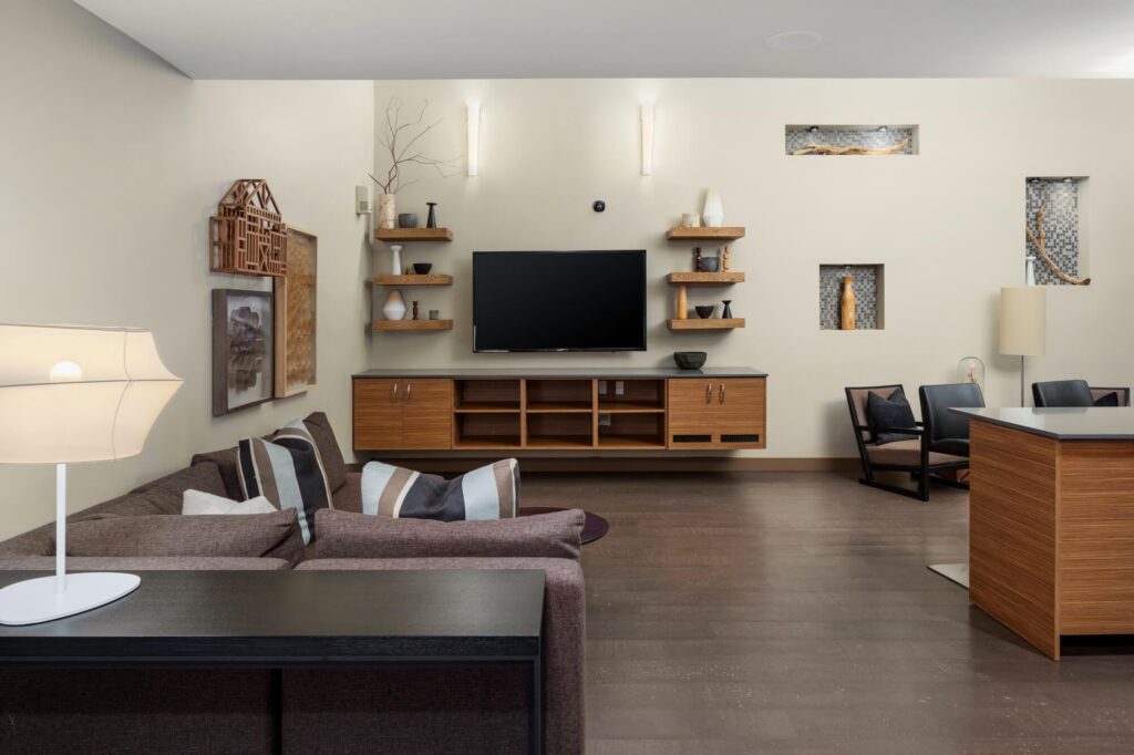 Modern living room with a brown sectional sofa, wall-mounted TV, wooden shelves and cabinets, decorative art pieces, two black chairs, and neutral-colored walls with built-in display niches.