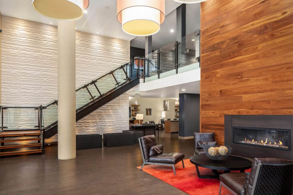 Modern hotel lobby with a wavy textured wall, floating staircase, wood-paneled wall with a fireplace, and a seating area featuring dark chairs, a round table, and a red rug under large hanging lights.