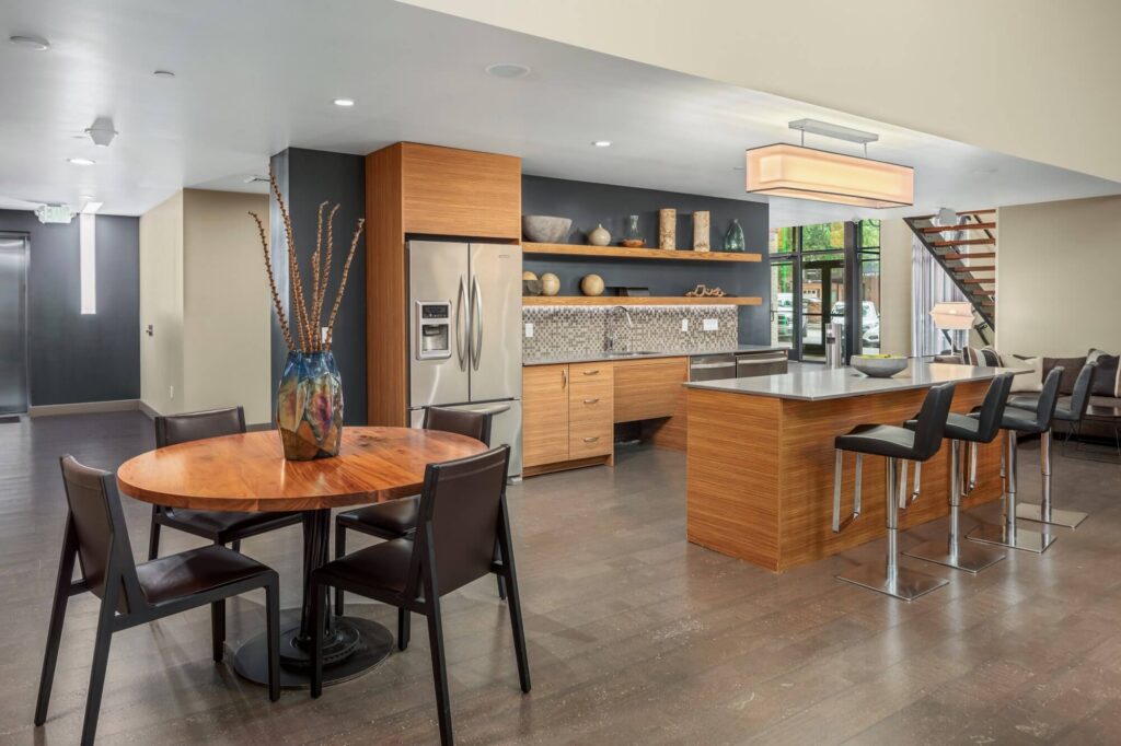 Modern open-concept kitchen and dining area with wood cabinets, stainless steel appliances, island with barstools, round dining table, leather chairs, decorative vase, and large windows.