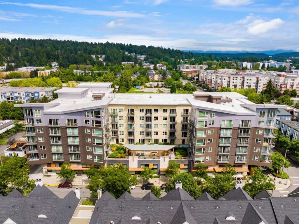 Aerial view of a modern mid-rise apartment building with balconies and green landscaping, surrounded by trees and other residential buildings on a sunny day. Forested hills are visible in the background.