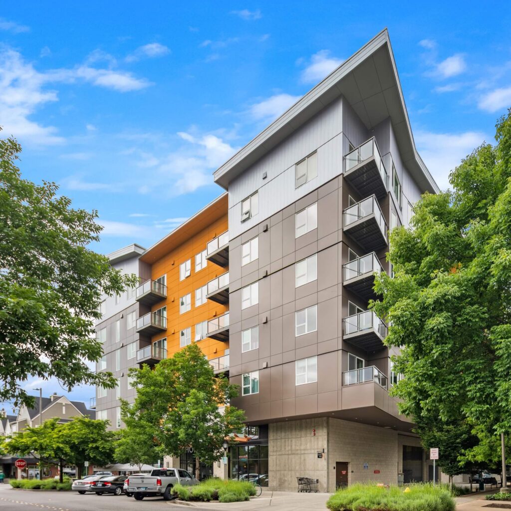 Modern mid-rise apartment building with balconies and a mix of gray, white, and orange panels, surrounded by green trees and parked cars, under a bright blue sky with scattered clouds.