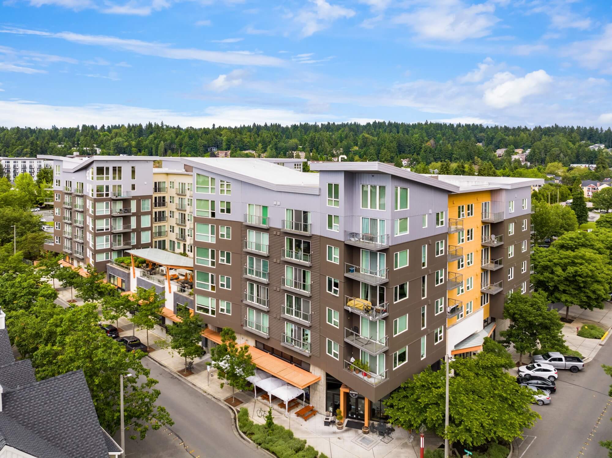 A modern mid-rise apartment building with large windows and balconies sits on a tree-lined street corner, surrounded by greenery under a partly cloudy blue sky.