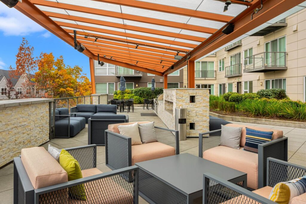 Outdoor patio with modern lounge seating, a covered pergola, cushioned chairs, a stone fireplace, and a dining area. Surrounded by greenery and apartment buildings, with autumn trees in the background.
