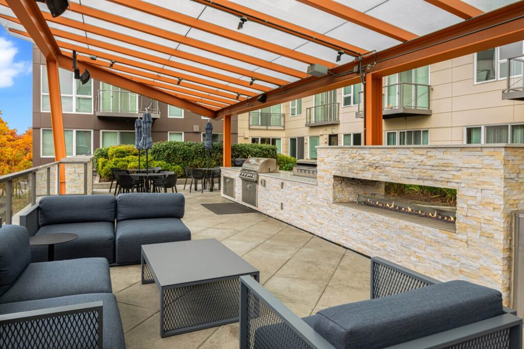 Outdoor patio area with modern seating, tables, a barbecue grill, and a stone fireplace under a pergola, surrounded by apartment buildings and greenery.