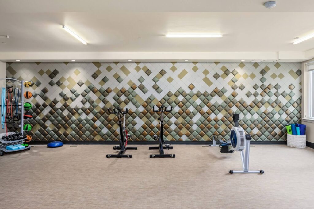 A modern gym room with a patterned tile accent wall, two stationary bikes, a rowing machine, and racks of colorful exercise equipment neatly organized along the side. The room is well-lit with overhead lights.