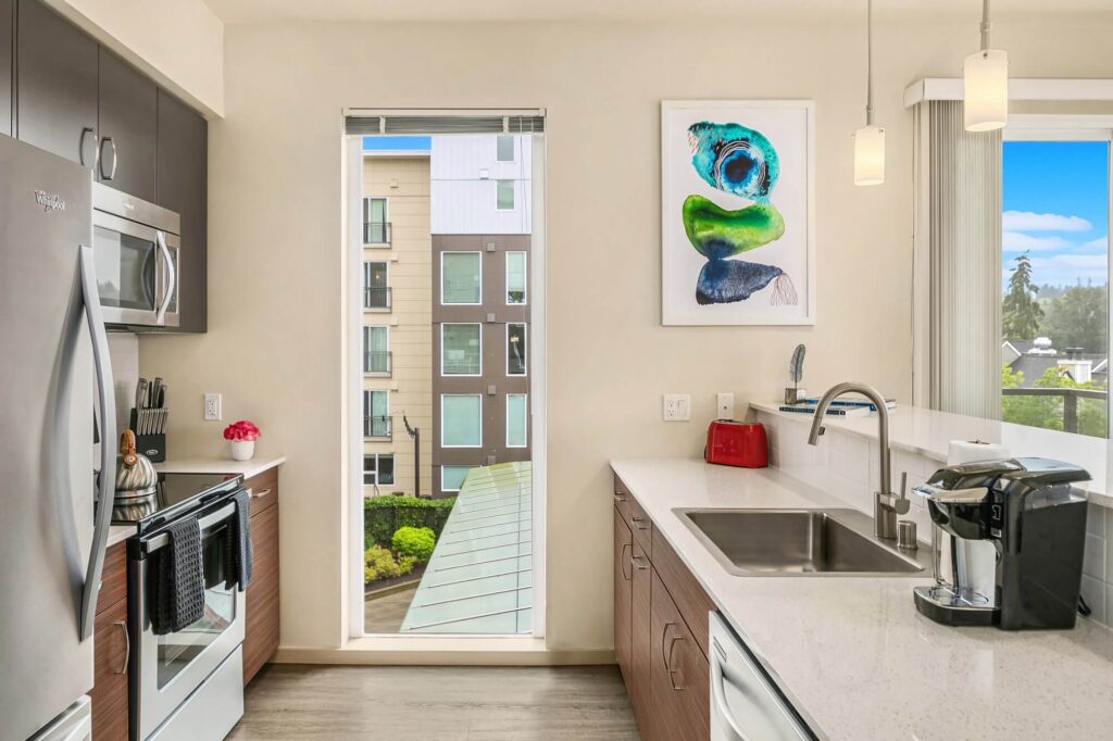 Modern kitchen with stainless steel appliances, white countertops, a coffee maker, red toaster, abstract wall art, and a large window overlooking contemporary apartment buildings and greenery.