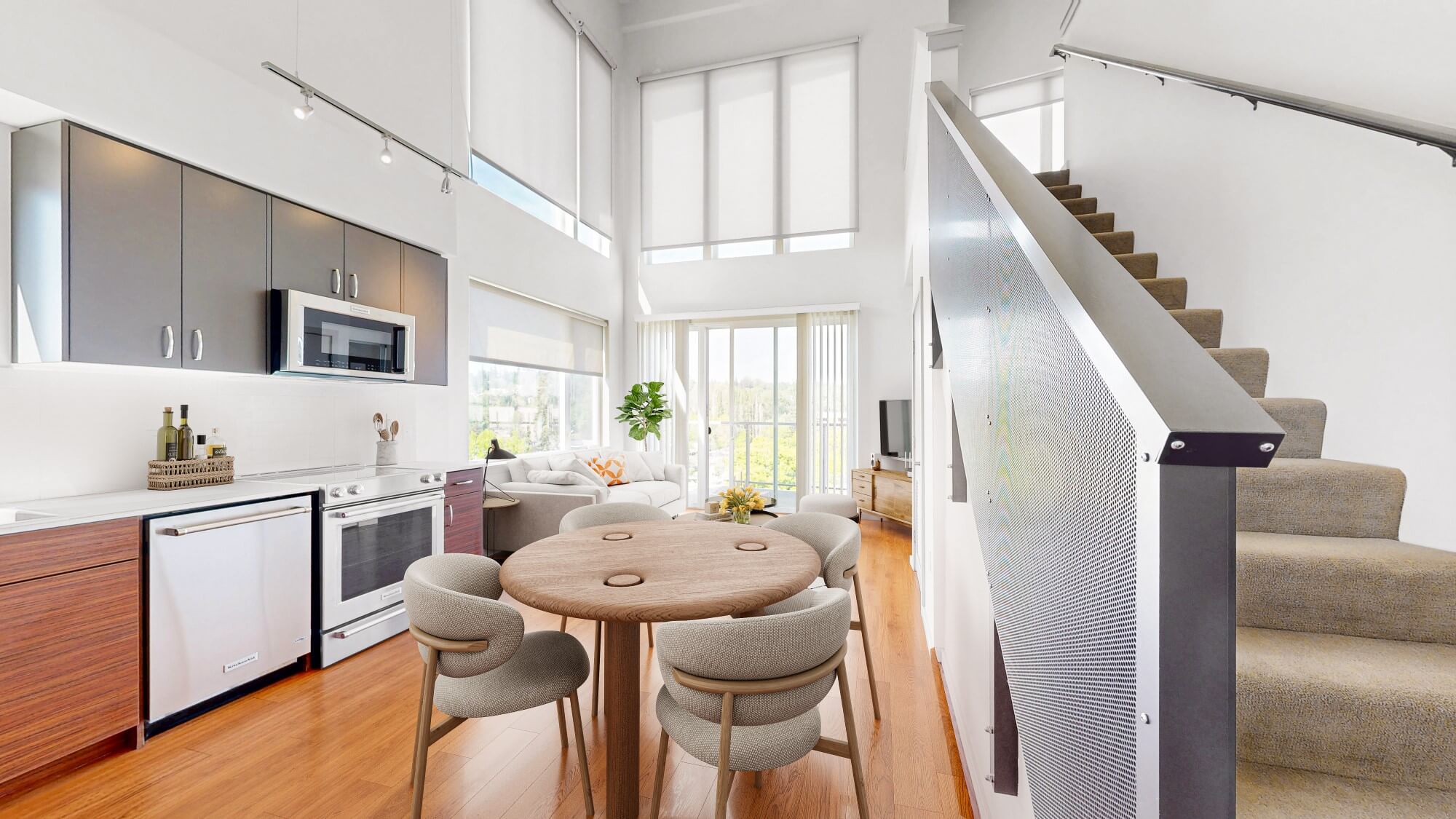 Bright modern apartment interior with an open kitchen, round dining table and chairs, cozy living area with a sofa and TV, large windows, wood floors, and a carpeted staircase leading to an upper floor.