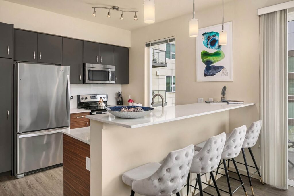 Modern kitchen with dark cabinets, stainless steel appliances, a white island counter with four tufted barstools, pendant lights, large windows, and abstract artwork on the wall.