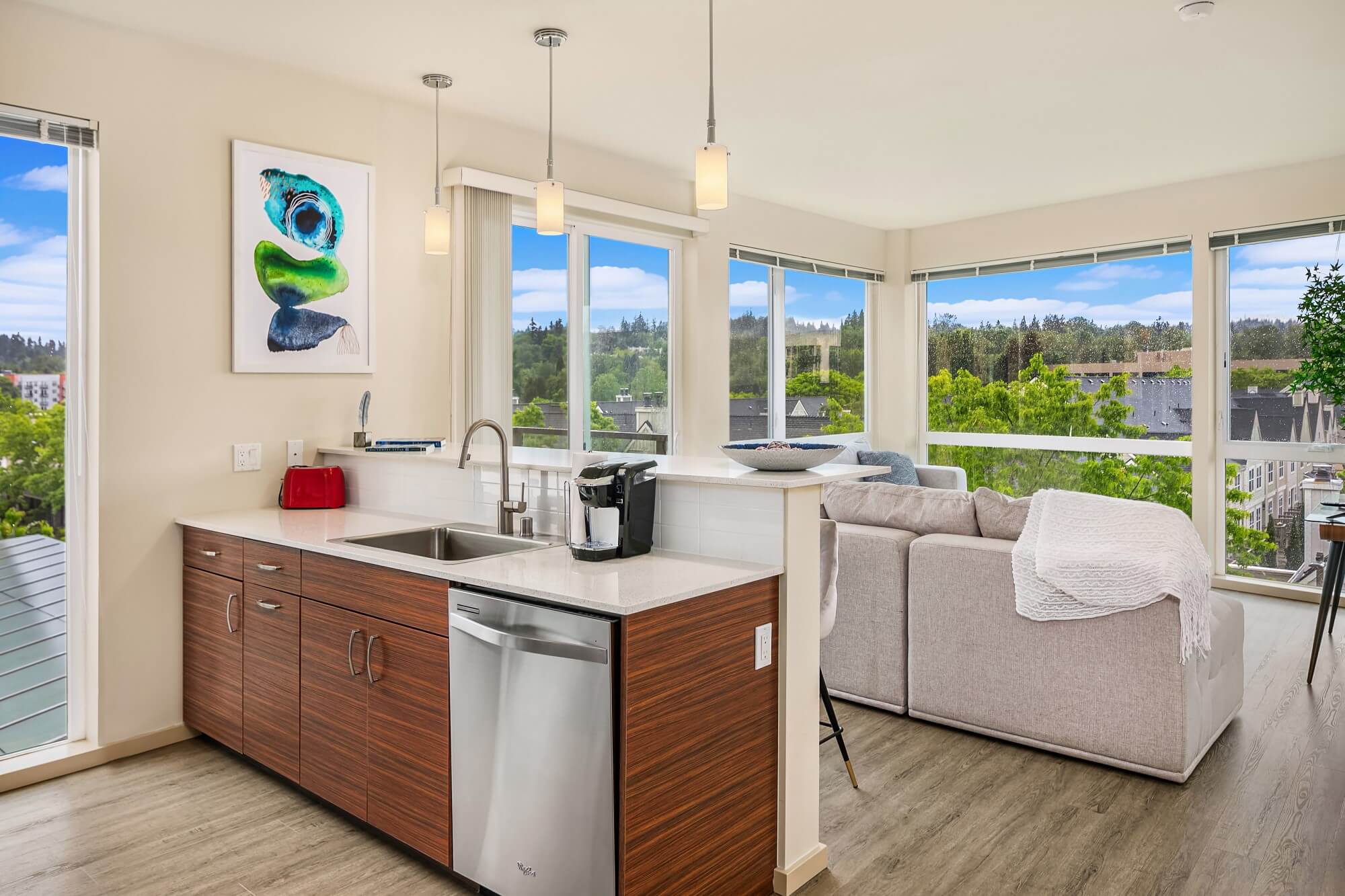 Modern open-concept kitchen and living room with wood cabinets, stainless steel dishwasher, and sink. Large windows offer views of trees and blue sky. A sofa and artwork complete the bright, airy space.
