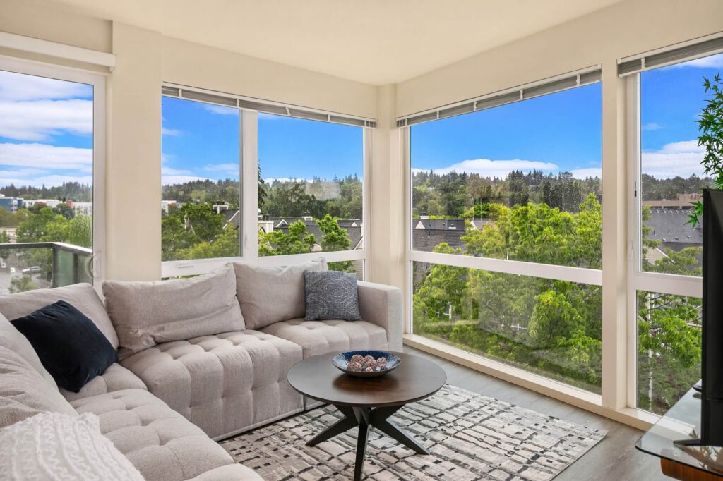 Bright living room with large corner windows, a light gray sectional sofa, a round coffee table with a decorative bowl, hardwood floors, and views of green trees and rooftops outside.