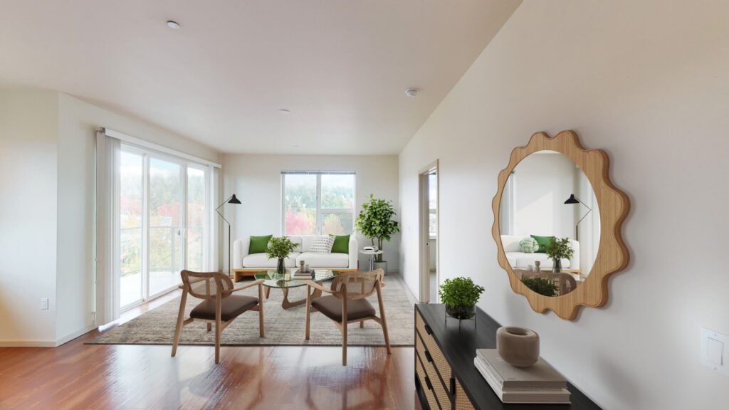 A bright, modern living room with large windows, wooden floors, a beige rug, two wooden chairs, a white sofa with green pillows, potted plants, and a dresser with a round mirror hanging above it.
