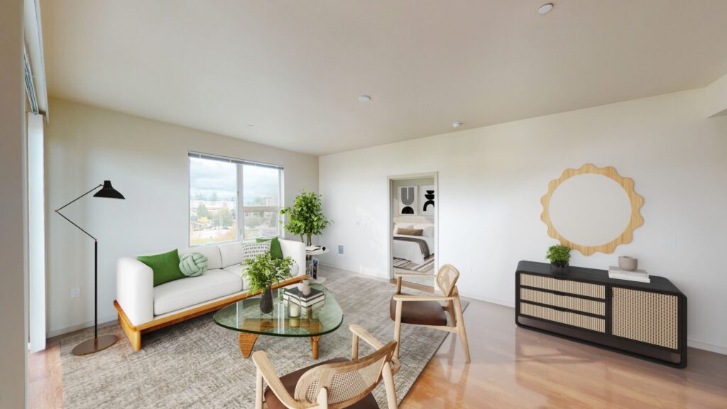A bright, modern living room with a white sofa, green pillows, round glass coffee table, two wooden chairs, a black floor lamp, a potted plant, and a console with a round mirror; window and open doorway in the background.