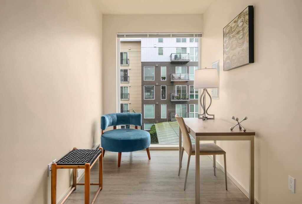 A small, modern room with light walls, featuring a blue accent chair, a wooden desk with a chair and lamp, a wall-mounted artwork, and a window overlooking an apartment building with balconies.