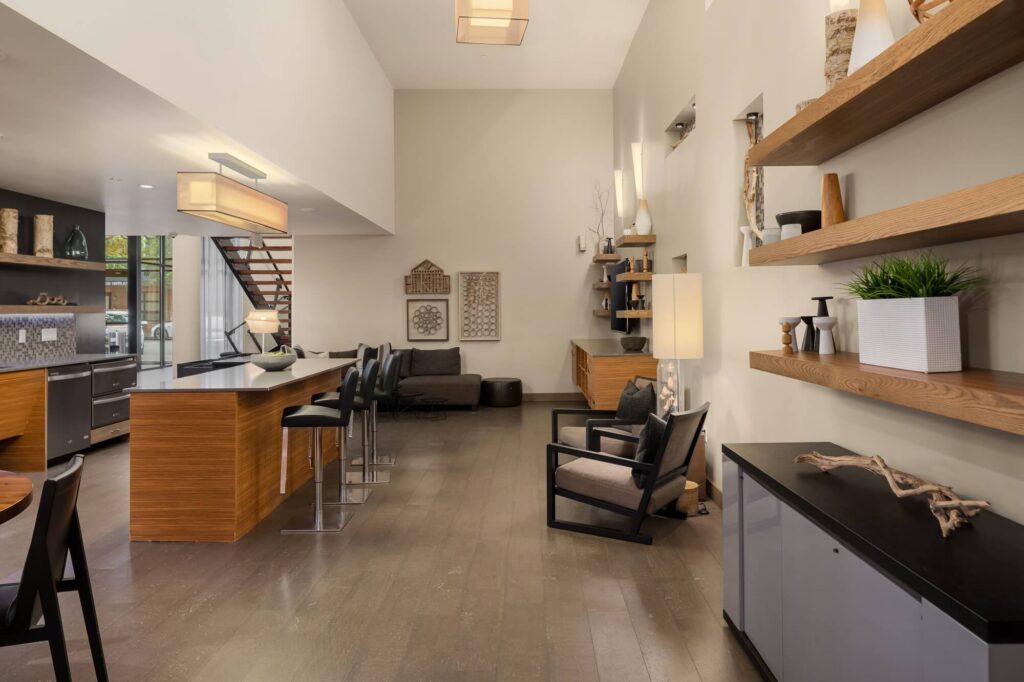 Modern open-plan kitchen and living area with wooden floors, island with bar stools, floating shelves with decor, gray sofa, armchair, large windows, and stairs leading upstairs. Neutral tones and contemporary design.